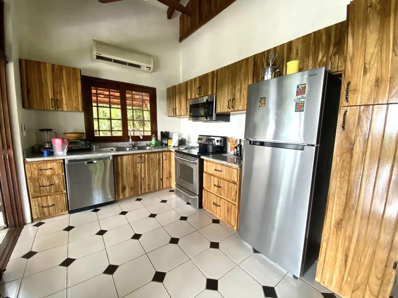 Casa Blue Wind · 16. This kitchen features rich natural wood cabinetry, a patterned tiled floor, stainless steel appliances, and a window above the sink offering views of tropical foliage and natural light, complemented by an overhead air conditioning unit.