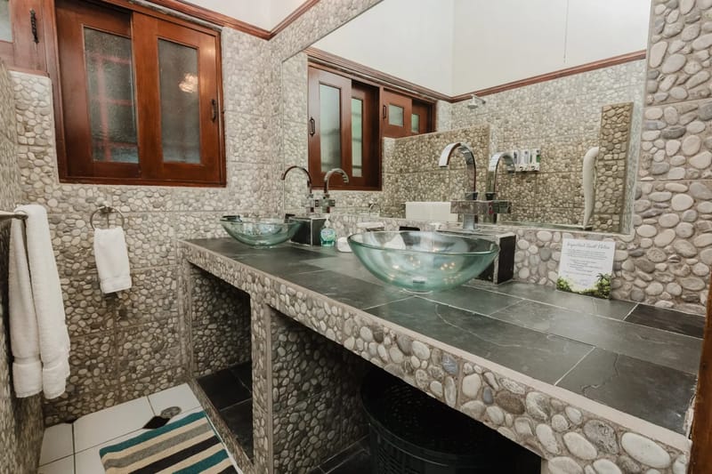 Casa Blue Wind · 18. The bathroom features pebble-tiled walls, a dark stone vanity with two glass vessel sinks, and a wooden-framed window allowing natural light.