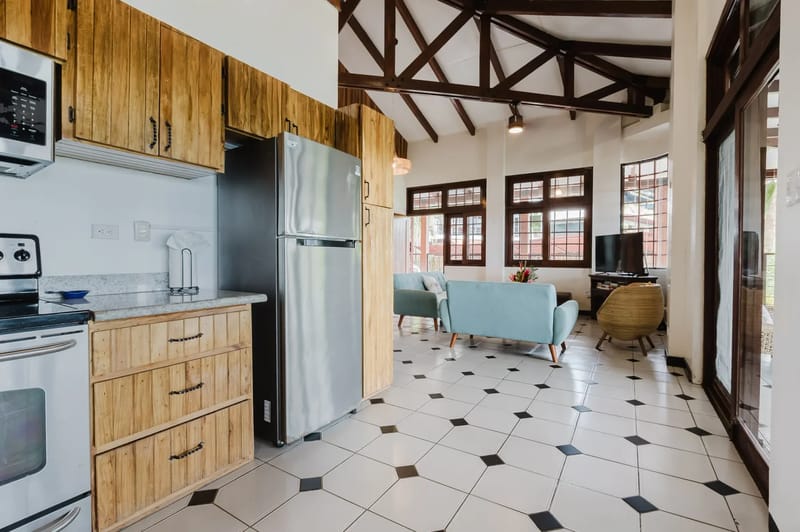 Casa Blue Wind · 29. The well-equipped kitchen, featuring natural wood cabinets and stainless steel appliances, opens to a bright living area with exposed beam ceilings, light blue seating, and grid-paned windows providing exterior views.