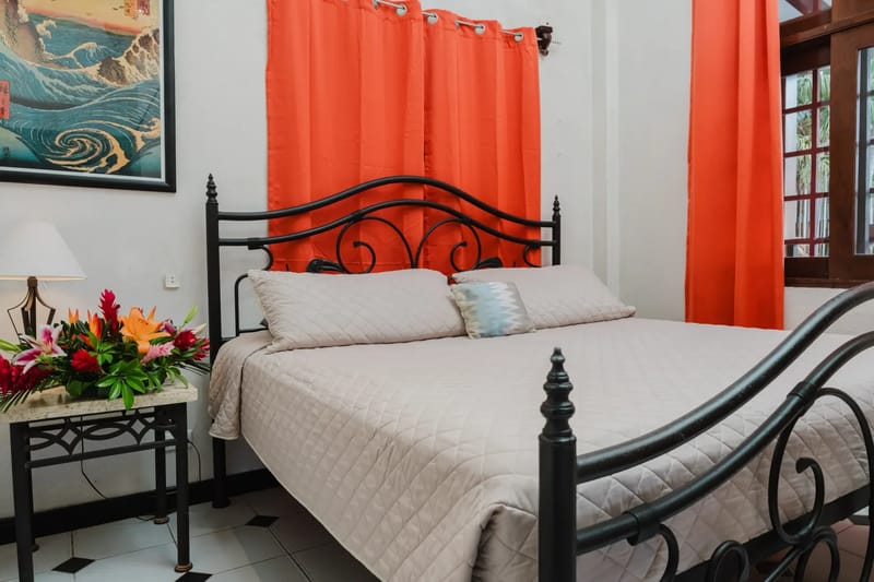 Casa Blue Wind · 23. This bedroom features a wrought iron bed, with bright orange curtains drawn over a window that offers filtered natural light and a view of tropical foliage.