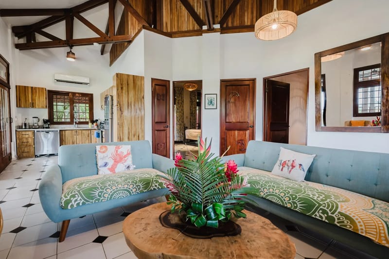 Casa Blue Wind · 10. This bright, open-plan living area and kitchen features a high ceiling with exposed wood beams and paneling, light tiled floors, two sofas, and natural wood cabinetry, with views of greenery through a kitchen window.