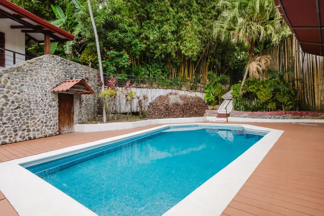 Casa Blue Wind · 11. A blue-tiled swimming pool surrounded by a brown deck is set against a backdrop of lush tropical foliage and a stone-faced structure with a red-roofed balcony.