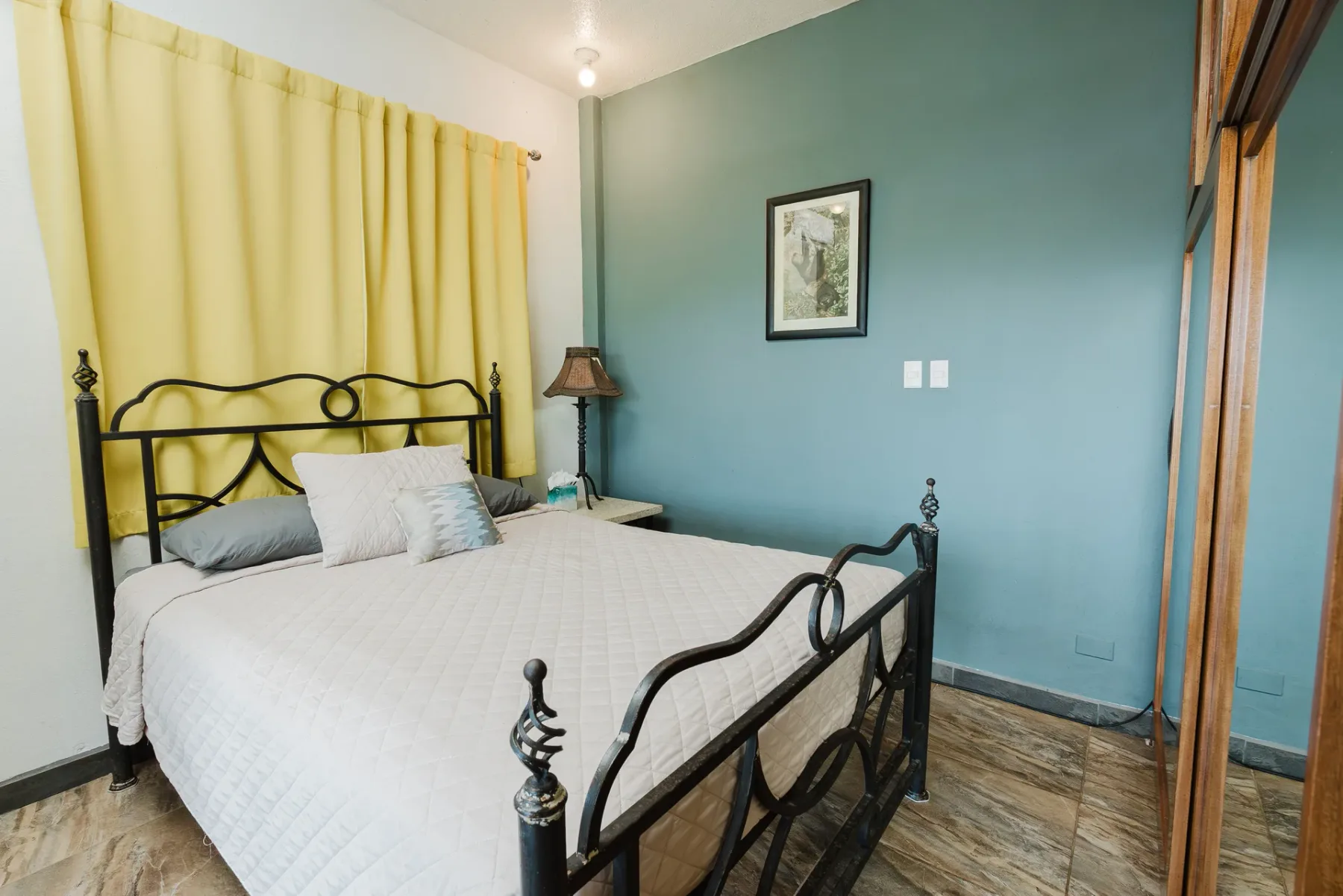 Casa Blue Wind · 24. This bedroom features a black wrought iron bed frame, a distinct teal accent wall, and light-colored tiled floors, with yellow curtains suggesting natural light from a window.