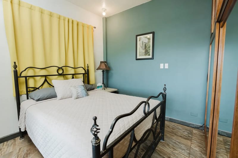 Casa Blue Wind · 24. This bedroom features a black wrought iron bed frame, a distinct teal accent wall, and light-colored tiled floors, with yellow curtains suggesting natural light from a window.