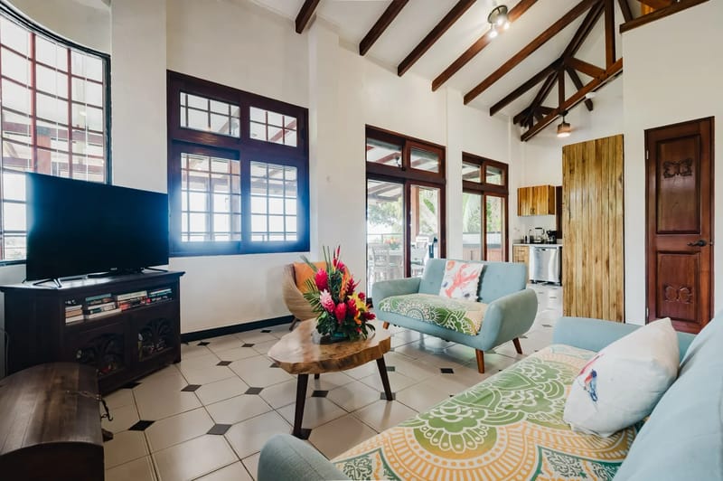 Casa Blue Wind · 9. The bright living room features high ceilings with exposed wooden beams, large windows, and sliding glass doors that open to an outdoor terrace.