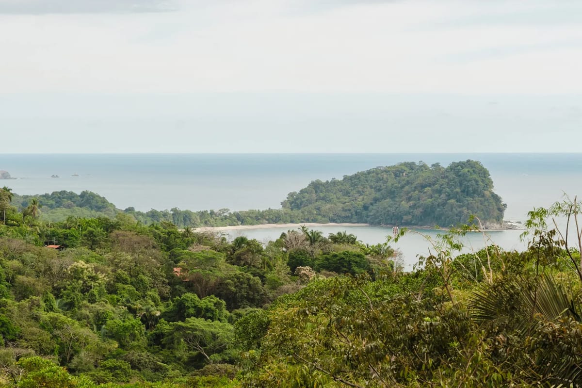 Casa Blue Wind · 1. A wide view showcases the ocean, a secluded bay with a sandy beach, a tree-covered island, and vast tropical jungle under an overcast sky.