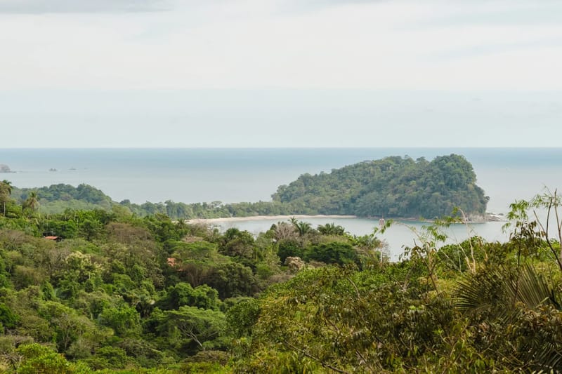 Casa Blue Wind · 1. A wide view showcases the ocean, a secluded bay with a sandy beach, a tree-covered island, and vast tropical jungle under an overcast sky.