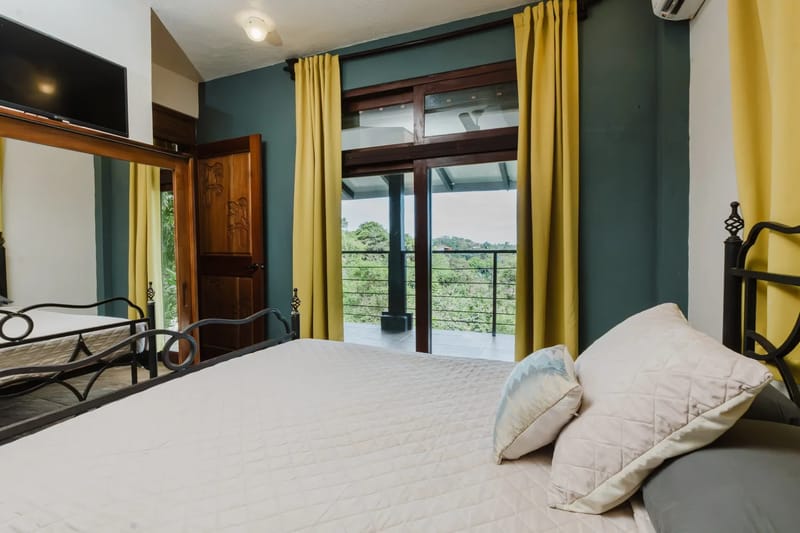 Casa Blue Wind · 31. This air-conditioned bedroom features a bed, a carved wooden door, and large glass doors that open to a private balcony overlooking a tropical landscape, providing ample natural light.