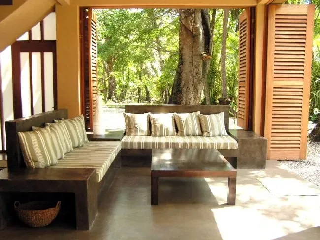 A covered outdoor seating area with cushioned benches and a low table overlooks leafy trees.