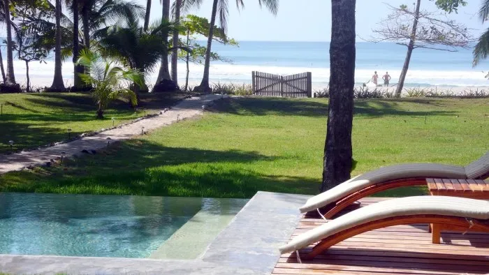 A swimming pool and wooden loungers overlook a grassy lawn and beach.