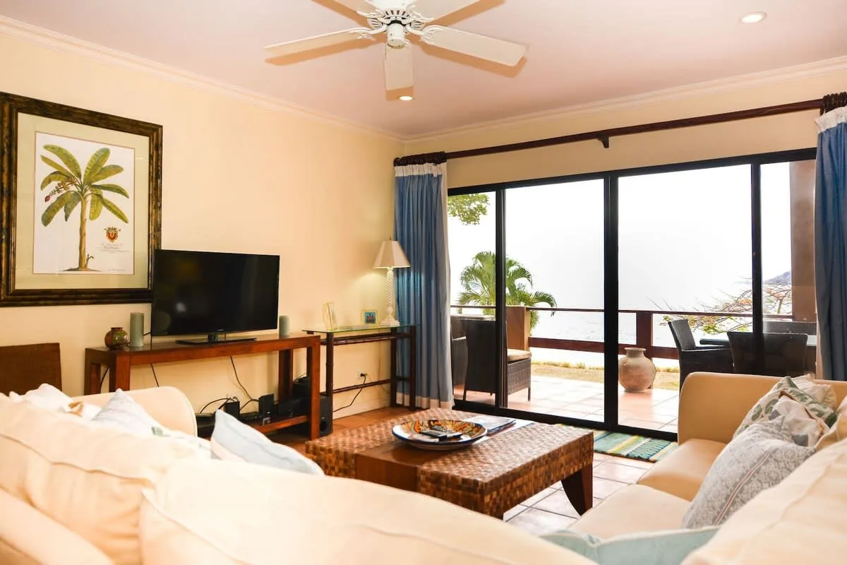 A bright living room with seating, a television, and large windows opening onto a patio.