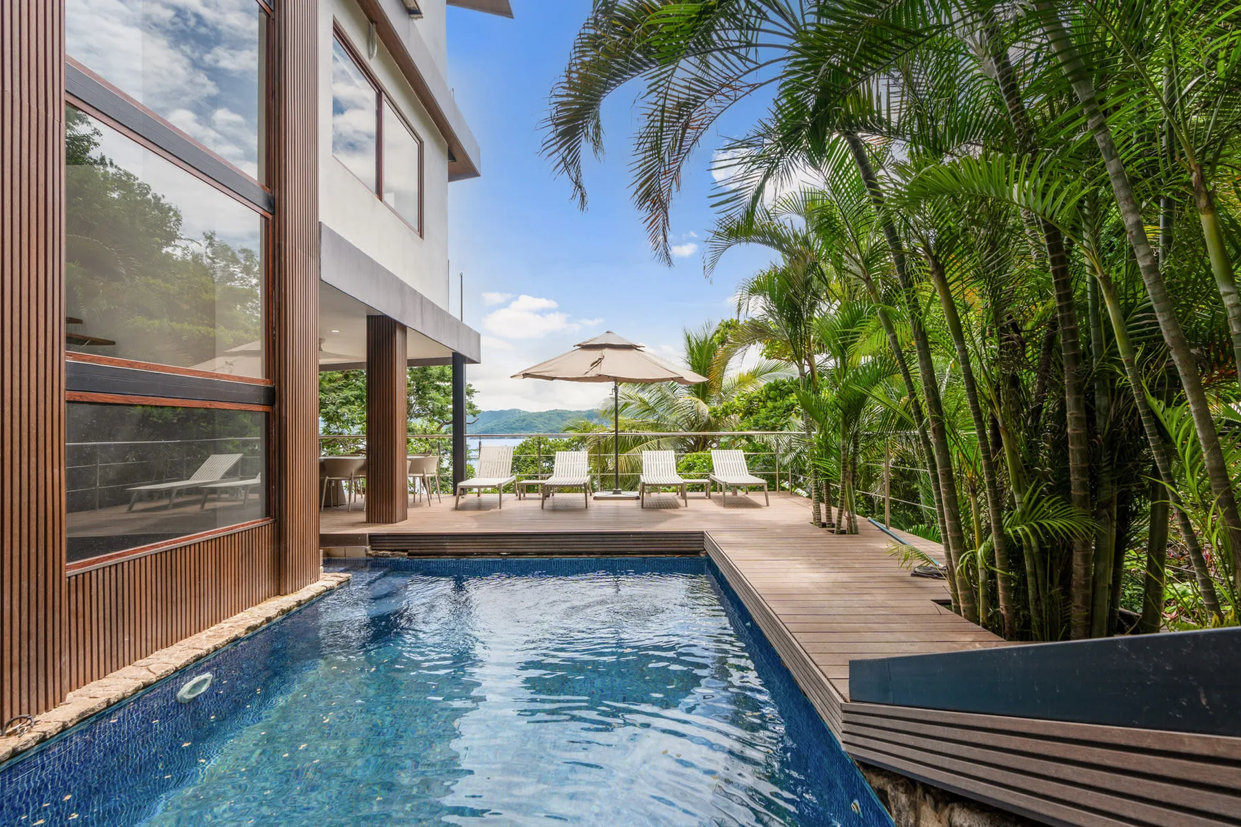 An outdoor swimming pool beside a wooden deck, lounge seating, and tropical palms.