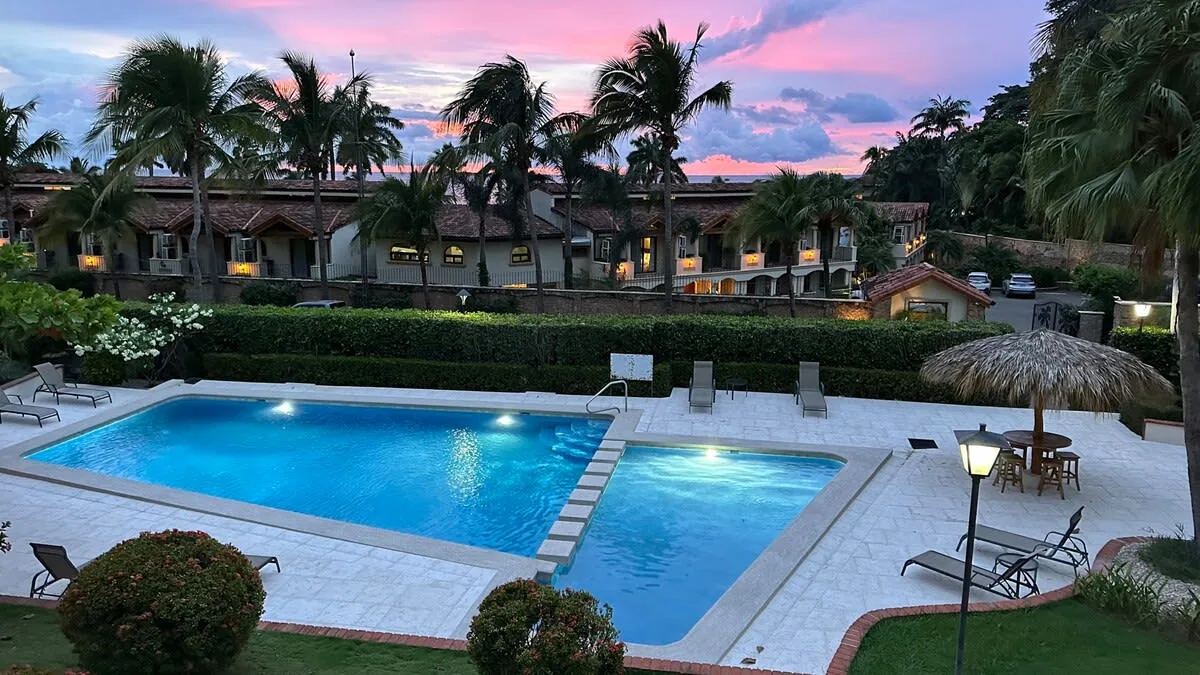 An outdoor swimming pool sits beside a smaller pool, with palms and buildings beyond.