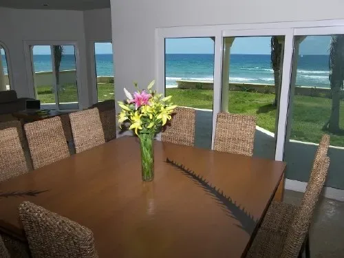 Una mesa de comedor con asientos tejidos está frente a grandes ventanas con vista al mar.