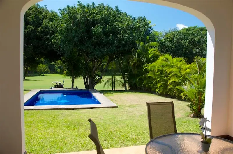 A patio overlooks a swimming pool and a green garden framed by trees.