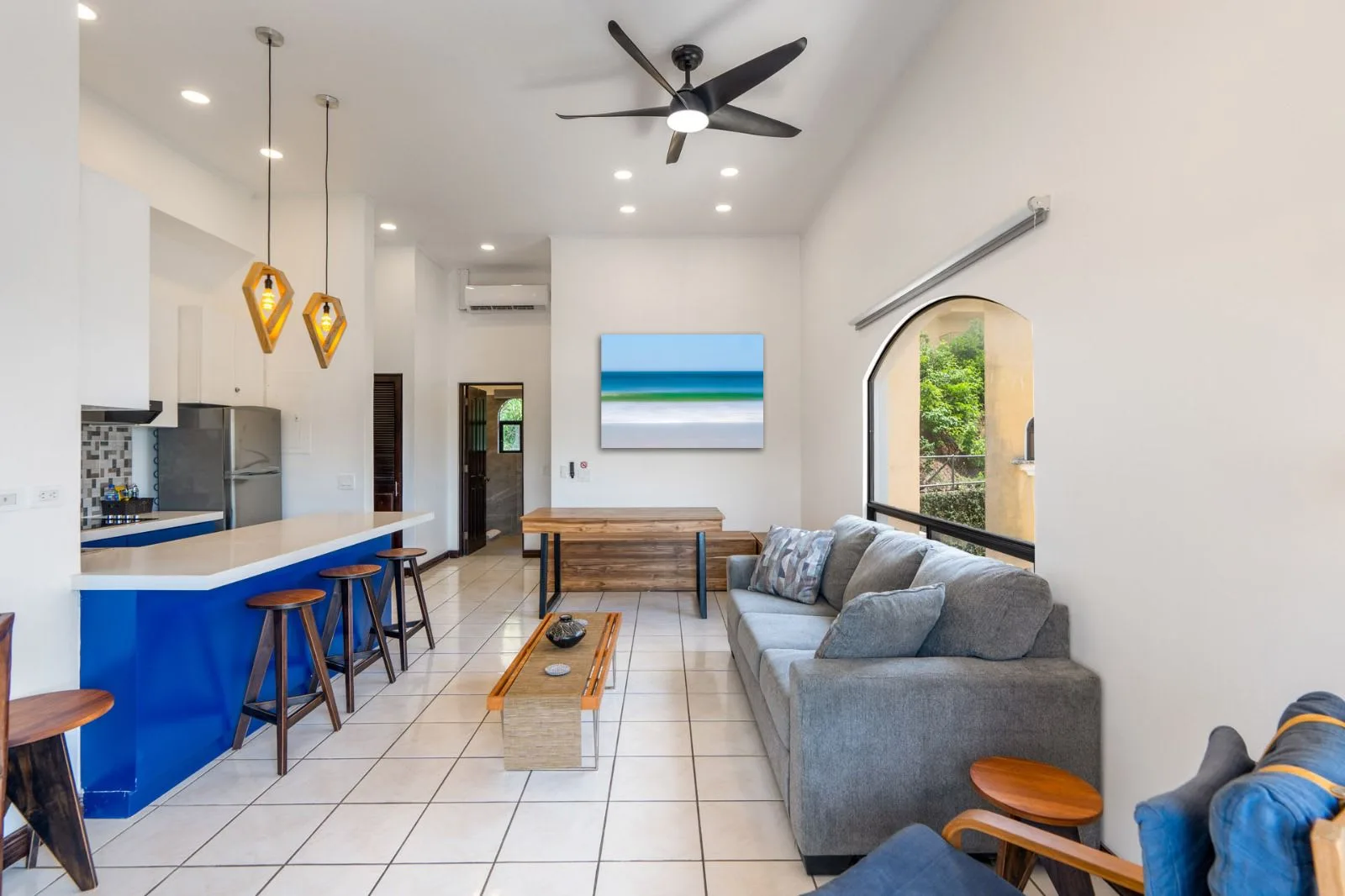 A bright living room with a gray seating area, blue kitchen counter, and ceiling fan.