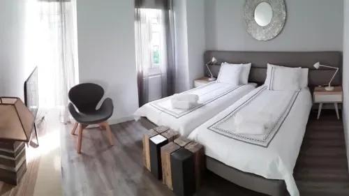 Un dormitorio luminoso con dos camas, un cabecero oscuro y una silla junto a la ventana.
