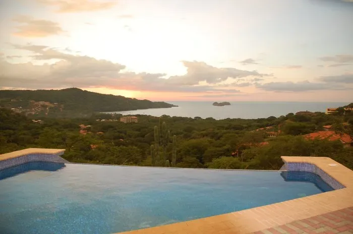 Una piscina infinita da a una costa verde y al océano al atardecer.