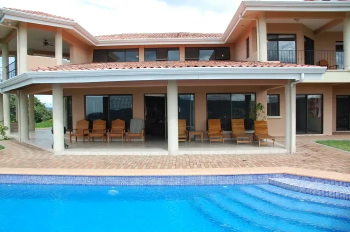 Una casa grande da a una piscina, con asientos al aire libre bajo un patio cubierto.