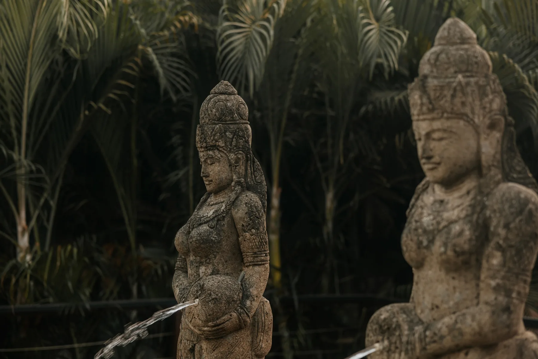 Dos estatuas de piedra desgastadas se alzan entre denso follaje tropical, junto a una pequeña fuente de agua.
