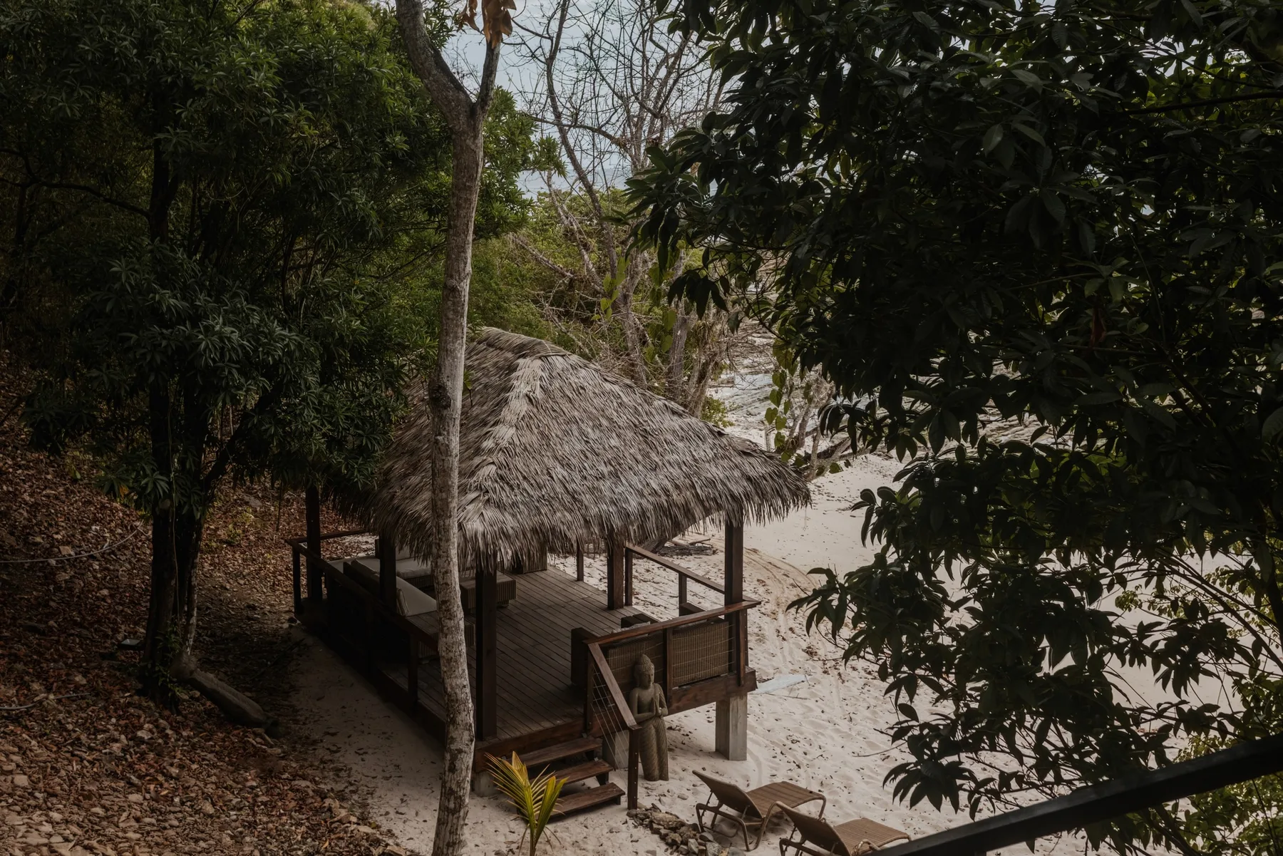 Una cabaña de madera con techo de paja está en una playa arenosa entre árboles frondosos.
