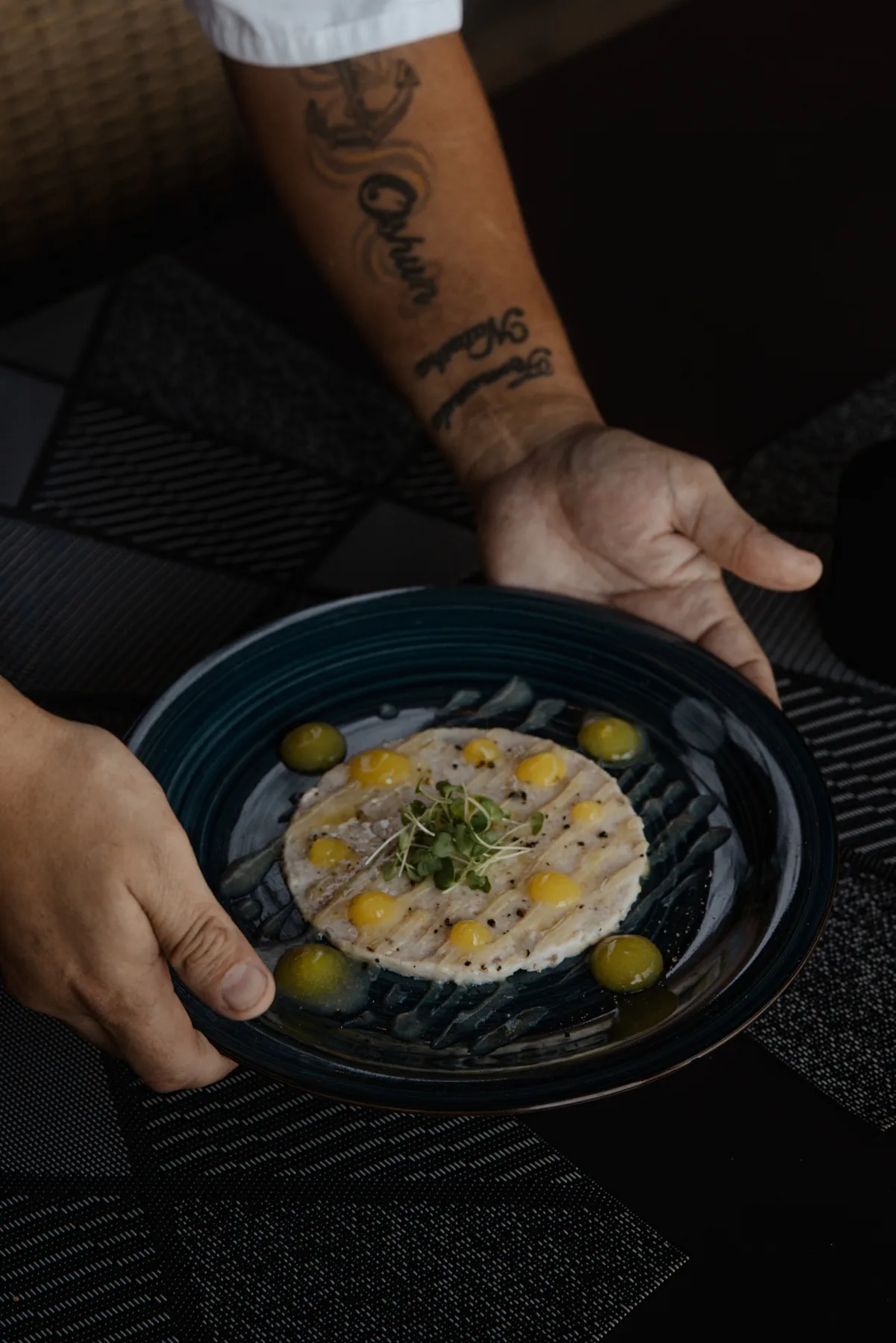Una persona presenta un plato oscuro con una preparación redonda cubierta de hierbas y rodeada de pequeñas aceitunas verdes.