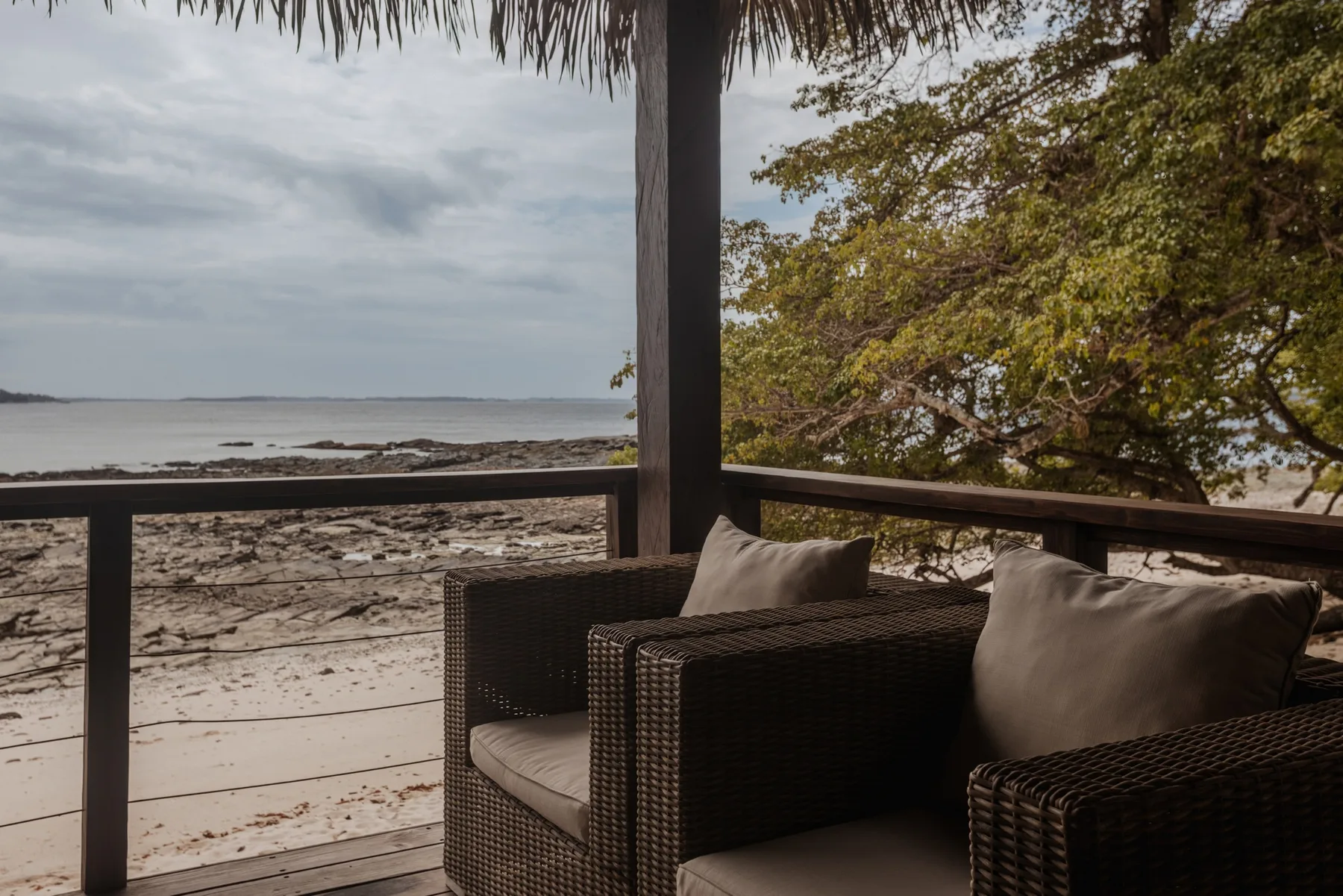 Un sillón de mimbre con cojines beige está en una terraza cubierta frente a una playa de arena y aguas tranquilas.