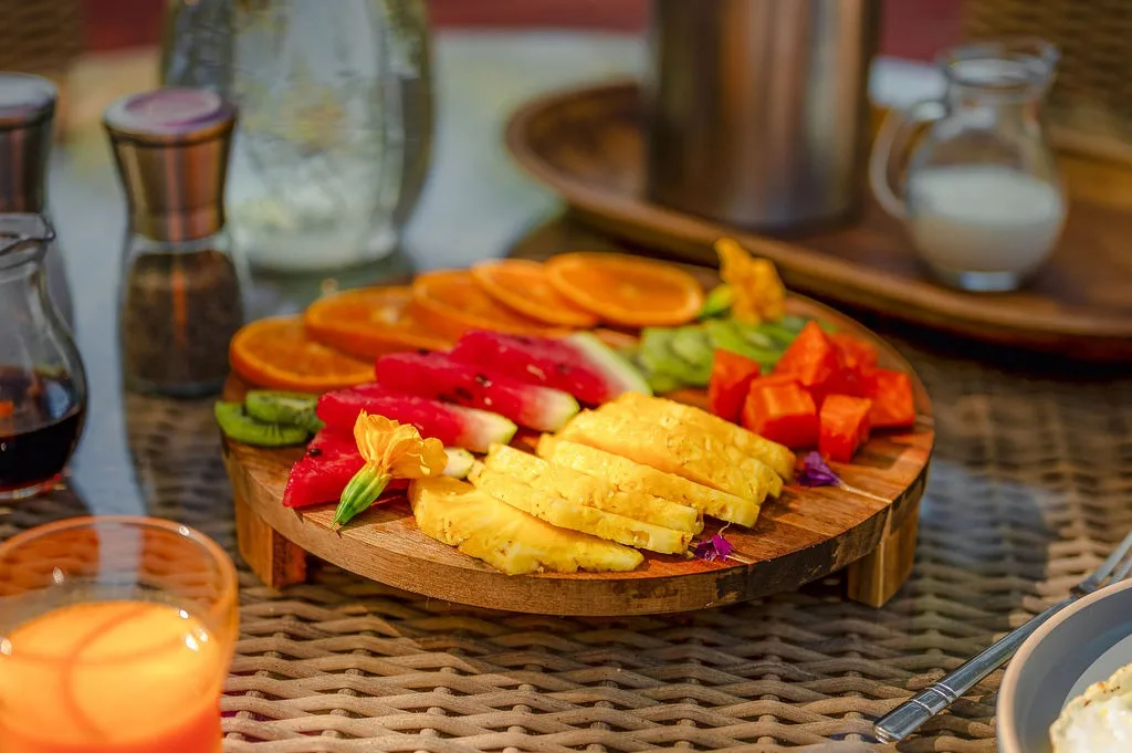 Una bandeja de madera con fruta y verduras cortadas está sobre una mesa trenzada, junto a condimentos y una bebida.