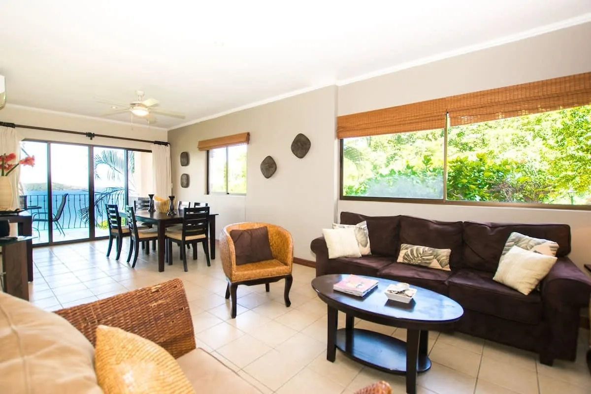 A living and dining area with dark seating beside large windows.