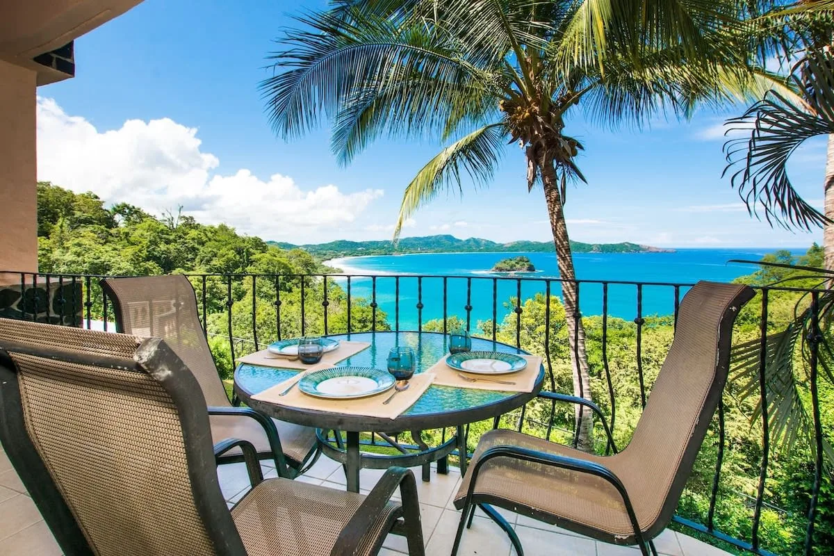 A balcony dining area overlooks palm trees and a bright blue bay.