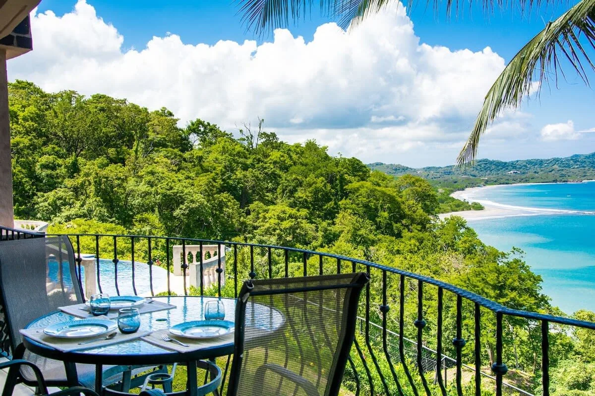 A balcony dining area overlooks lush greenery and a curving beach.