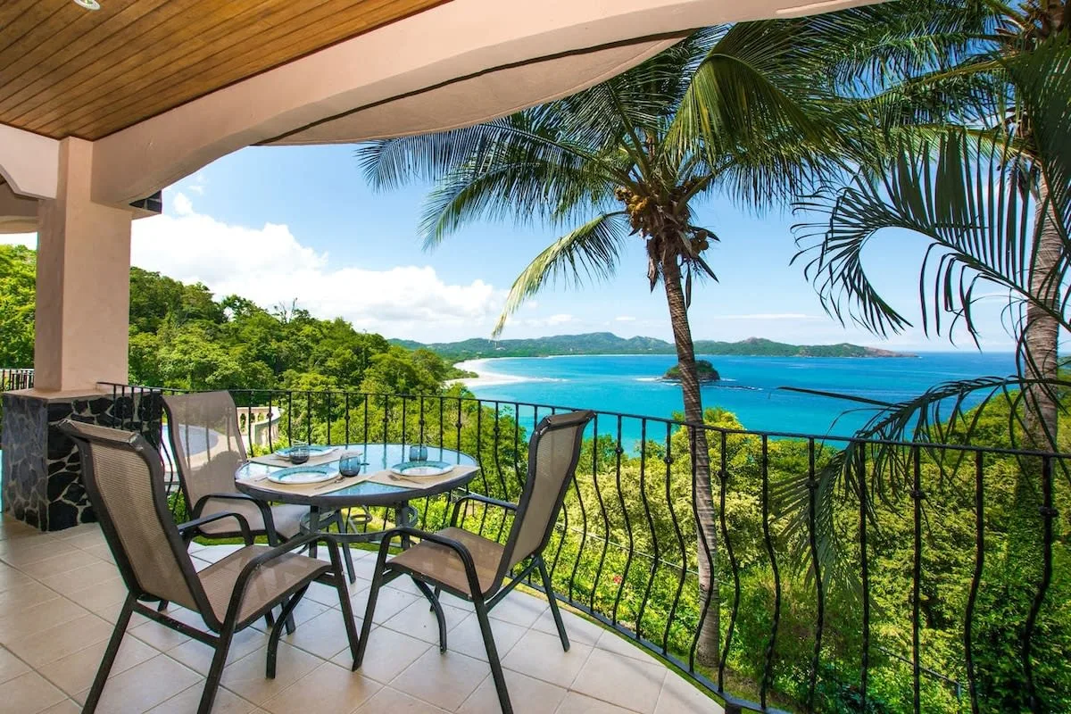 A covered balcony has outdoor seating overlooking palm trees and a blue ocean.