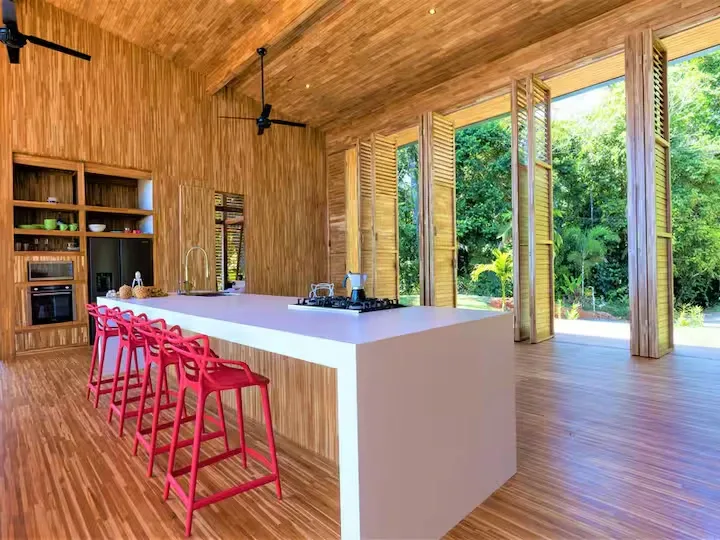 A wood-paneled kitchen and dining area opens onto lush greenery through wide windows.