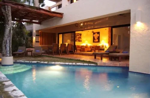 Una piscina exterior está frente a una sala iluminada con asientos.