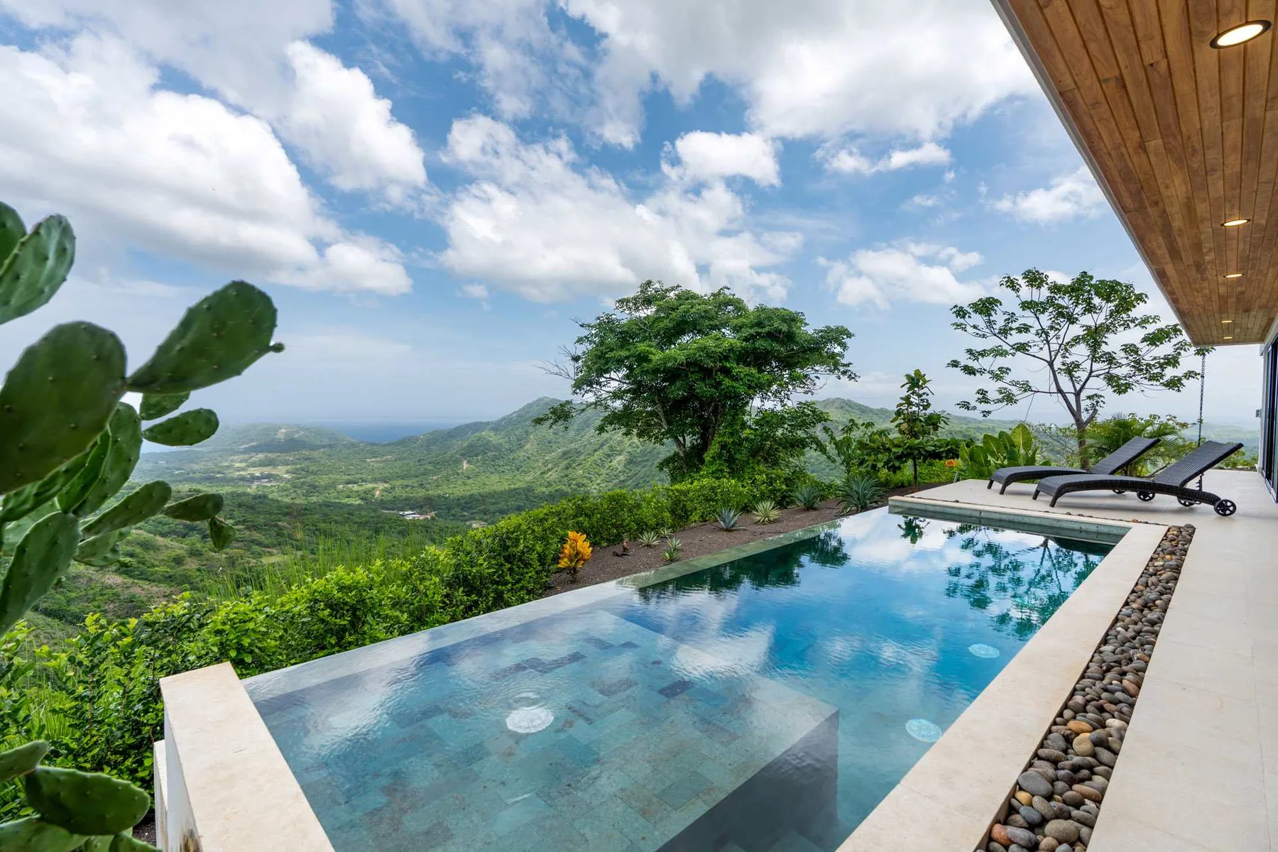 An outdoor swimming pool overlooks a green mountain landscape beneath a partly cloudy sky.