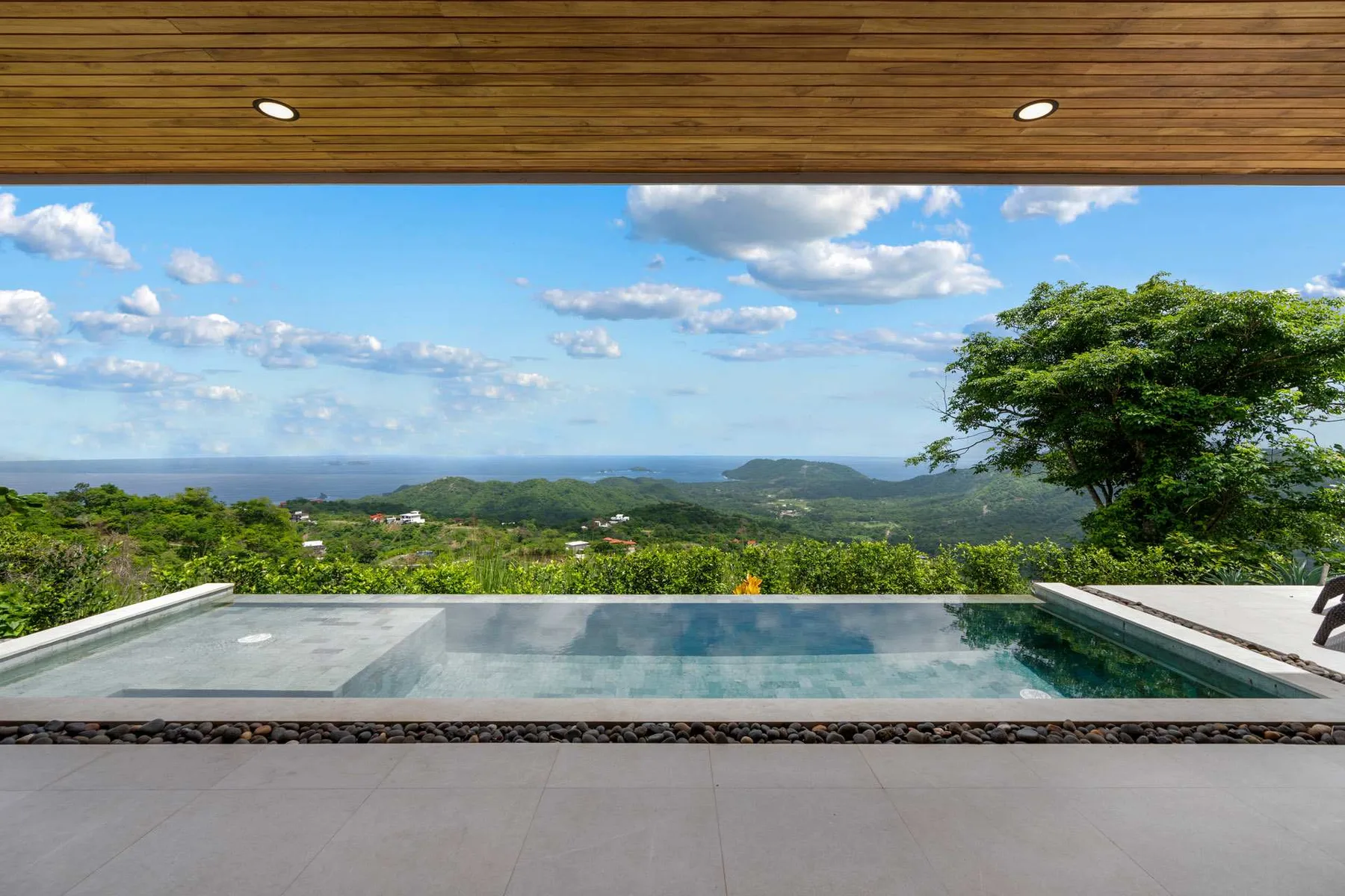 An infinity pool overlooks green hills and the ocean beneath a covered patio.