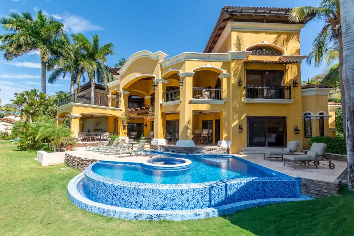 A large yellow villa stands beside a blue swimming pool and green lawn.
