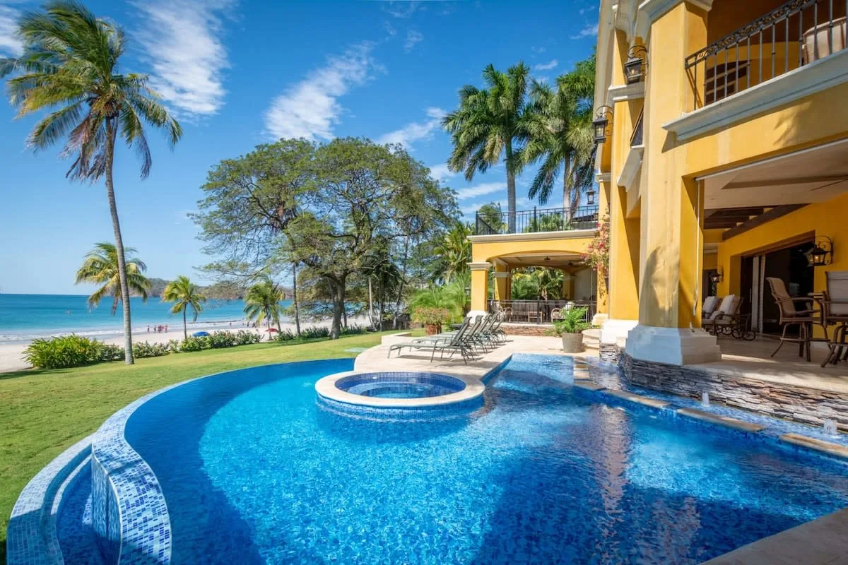 A swimming pool beside a yellow beachfront building, with palm trees and ocean views.
