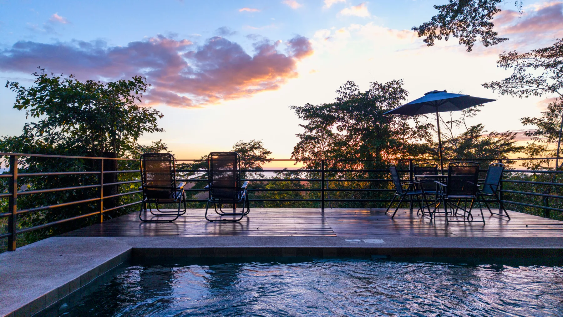 A swimming pool and deck seating overlook trees beneath a colorful sunset sky.