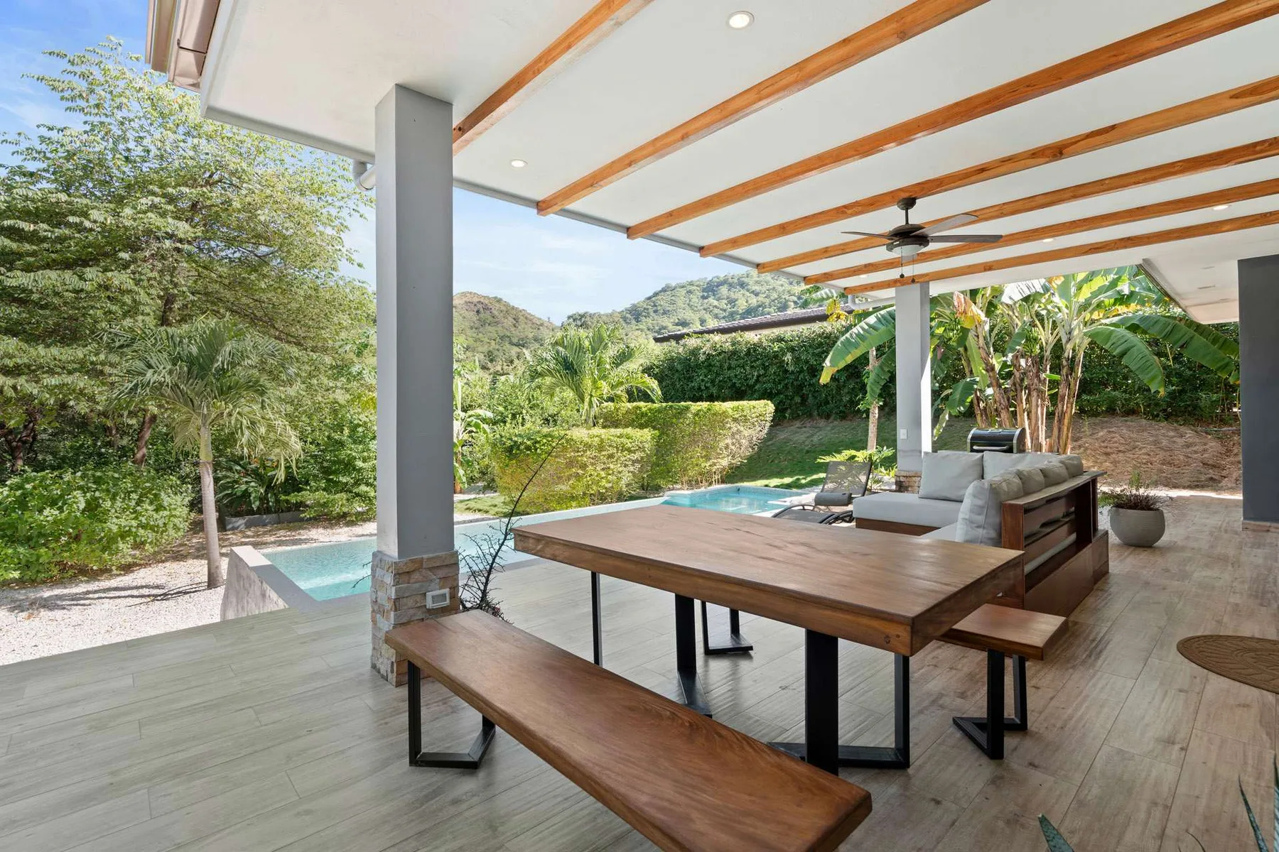 A covered patio with a wooden dining table, seating, and a pool beyond.
