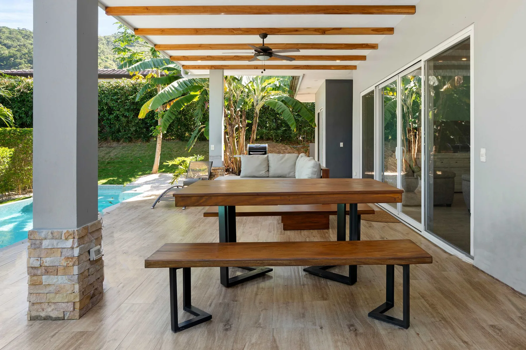 A covered patio features a wooden dining table and bench beside a swimming pool.