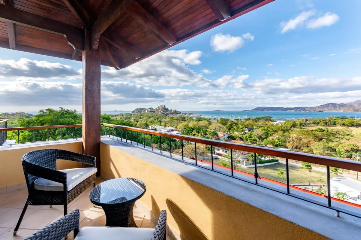 A covered balcony with seating overlooks greenery and a distant bay.