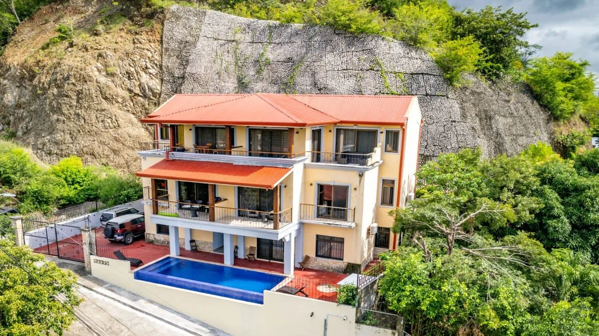 A multi-level house with balconies and a swimming pool sits beside a rocky hillside.