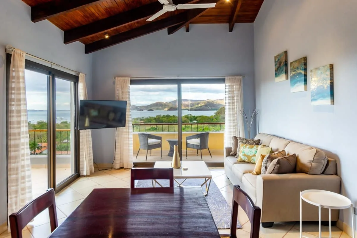 A bright living room opens onto a balcony with seating and a scenic view.