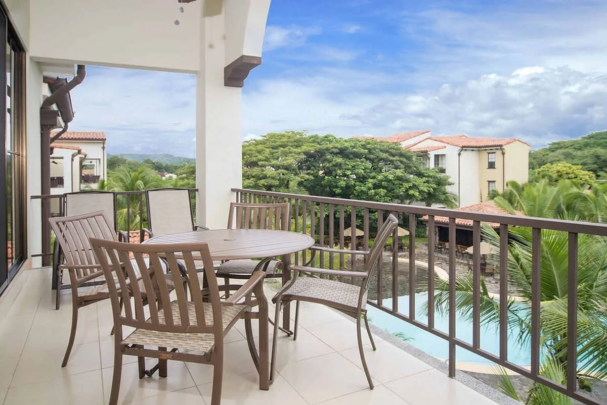 A tiled balcony has a wooden dining set beside a railing overlooking trees and a pool.