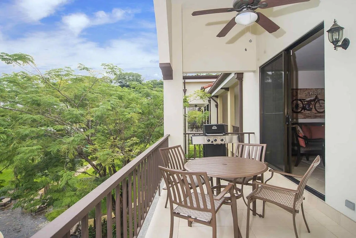 A balcony dining area with outdoor seating overlooking leafy trees.