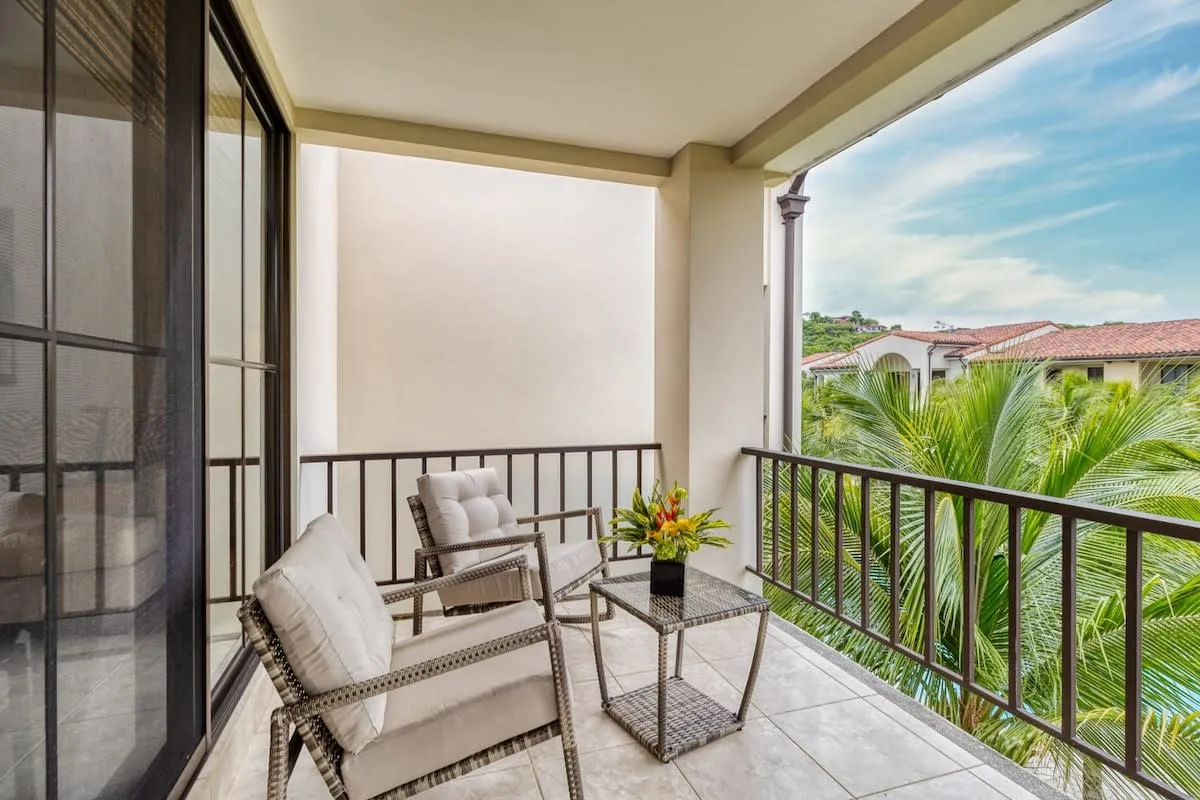 Un balcón cubierto tiene asientos y una mesa pequeña con vista a palmeras.
