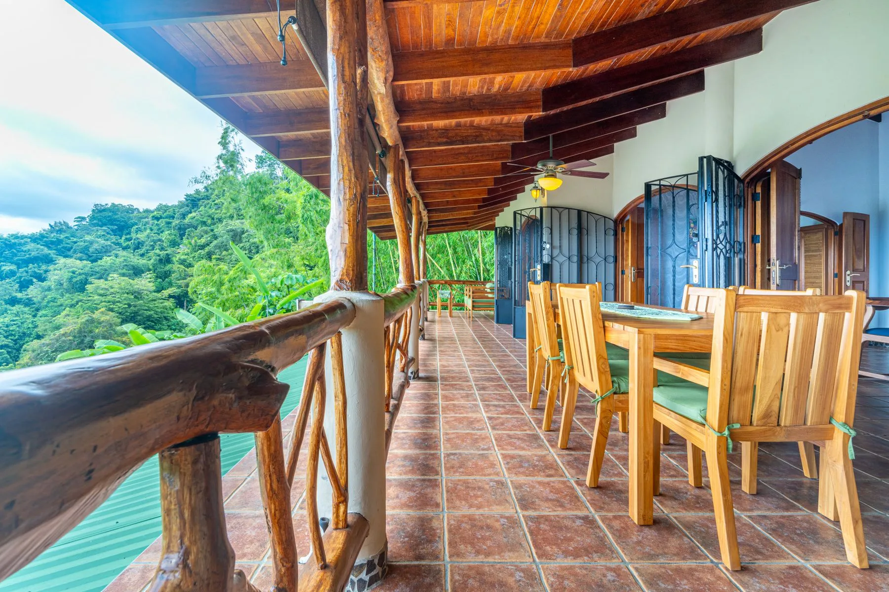 A covered wooden veranda has a dining table and chairs beside a green hillside.