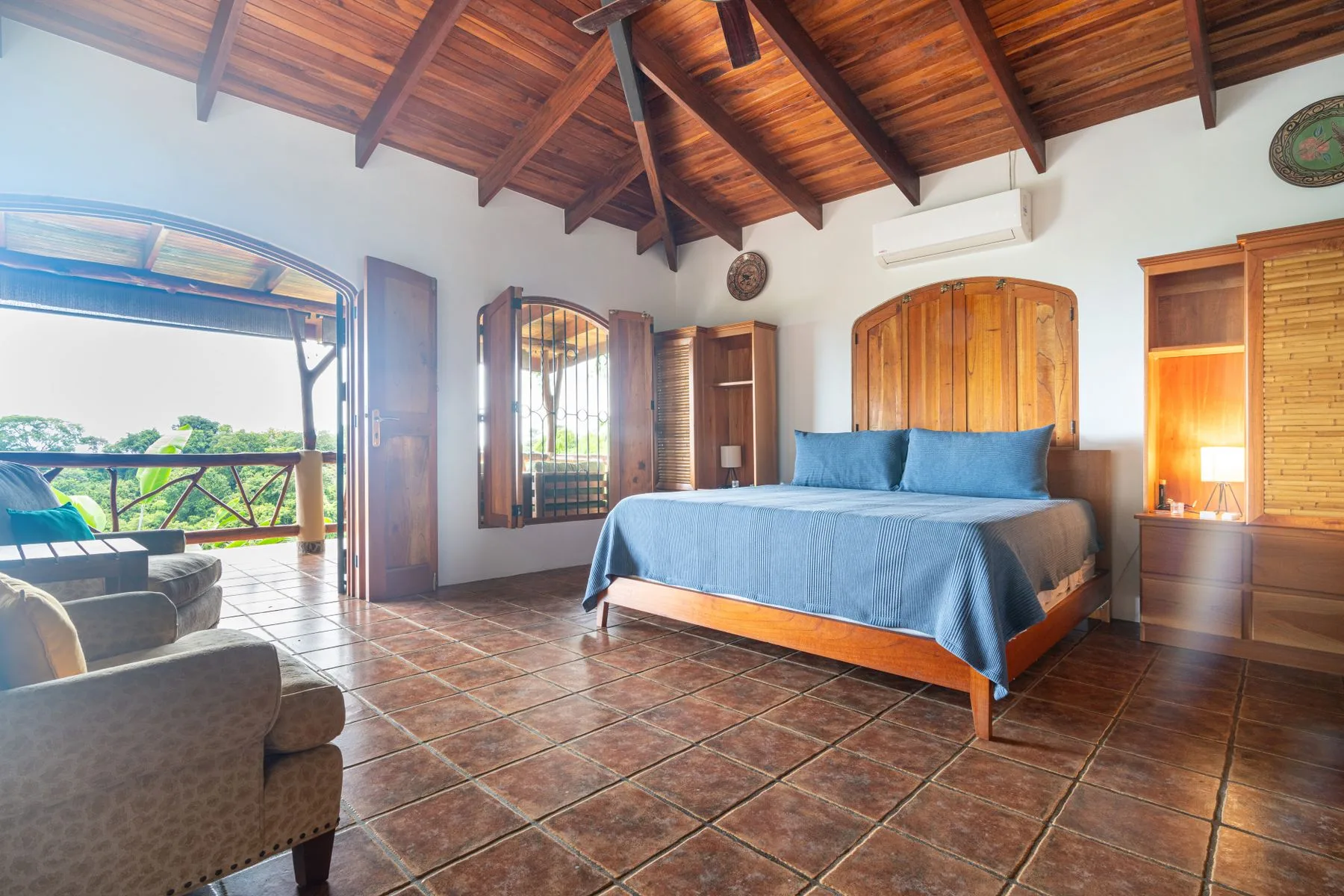 A bedroom with a blue-covered bed beneath a wooden ceiling, opening onto a balcony.