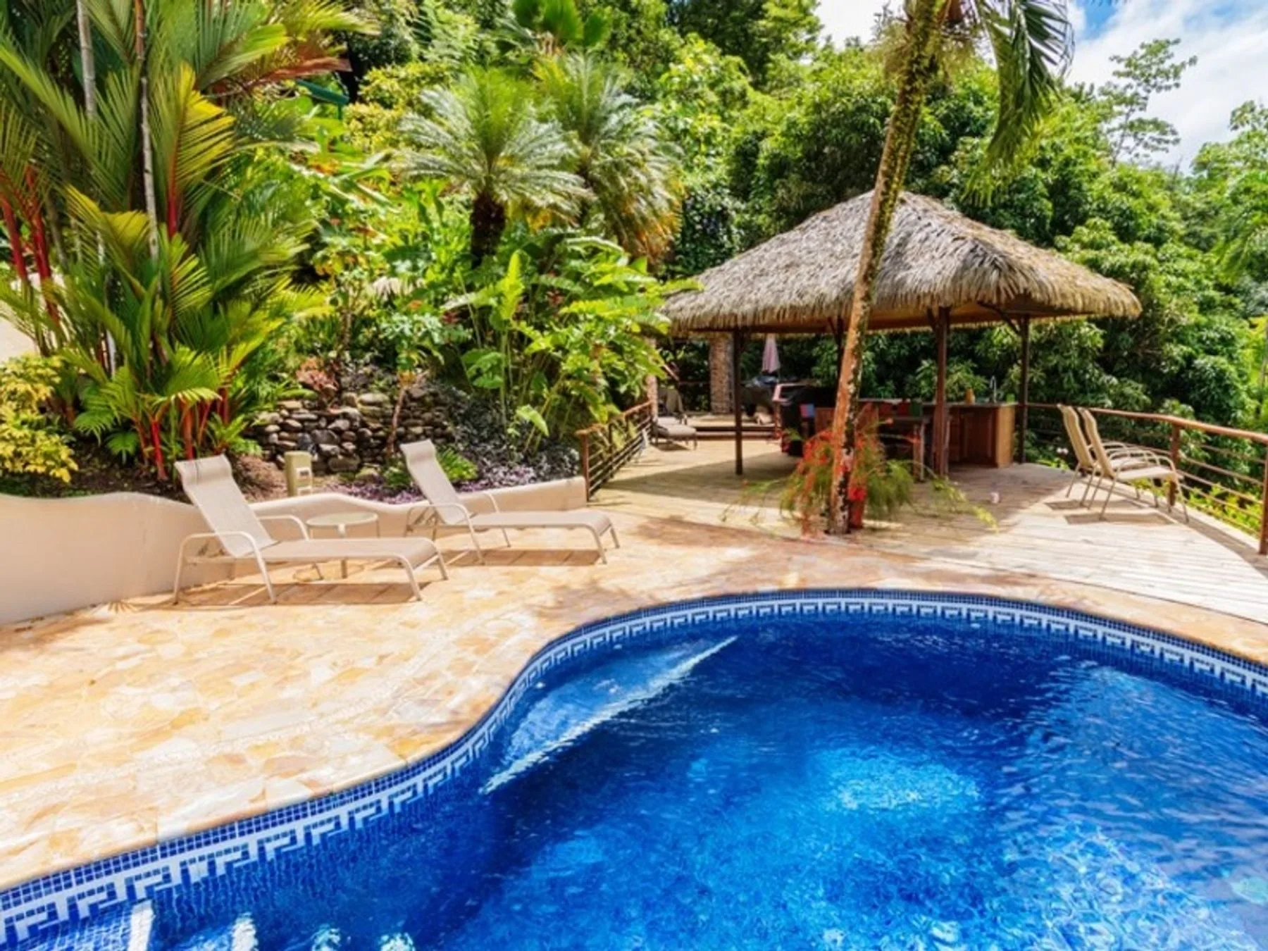 A swimming pool sits beside lounge seating and a thatched-roof pavilion amid dense greenery.